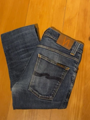 Blå jeans från Nudie Jeans - Säljer ett par klassiska blå jeans från Nudie Jeans med snygg tvätt och kontrastsömmar. Modellen är from Tim i stl W31 och L32. Jeansen är tillverkade i slitstark bomull och har en cool faded effekt på låren och knäna. W31 men sitter lite tajtare, typ som W30. Hör av er vid frågor✌️