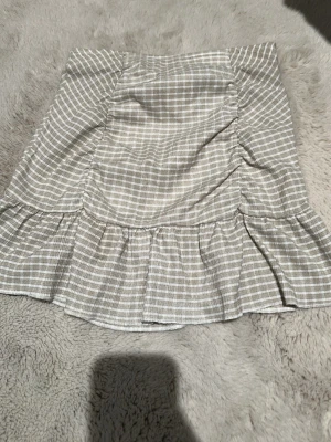 Beige och vit rutig kjol Gina Tricot - Kort kjol från Gina Tricot i beige och vitt rutigt mönster. Kjolen har volangkant nedtill och är tillverkad i ett lätt och mjukt tyg, perfekt för varmare dagar. Resår i midjan för skön passform och en söt, feminin look. Har inte använt den mycket ba någon gång sedan ba legat i garderoben.