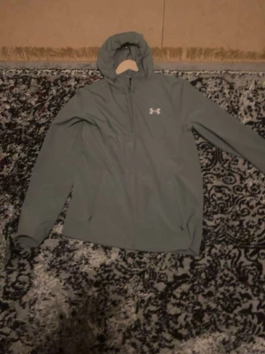 Grå vindjacka från Under Armour - Säljer en grå vindjacka från Under Armour med huva och dragkedja framtill. Jackan har en enkel och sportig design med logga på bröstet och långa ärmar. Perfekt för blåsiga dagar och passar till många olika outfits.