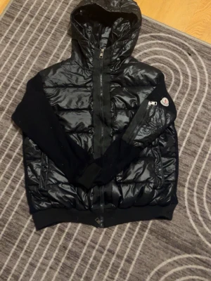 Svart Moncler pufferjacka med huva - Säljer en svart pufferjacka från Moncler med huva och dragkedja framtill. Jackan har quiltad kropp i glansigt material och stickade ärmar. Klassisk Moncler-logga på ena ärmen och praktisk ficka med dragkedja. Perfekt för kyliga dagar.