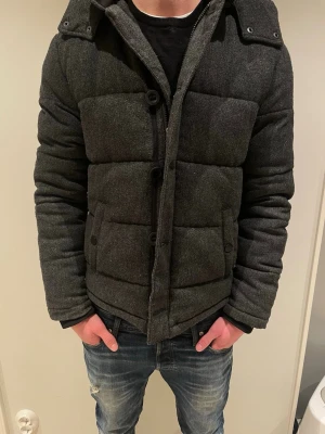 Mörkgrå pufferjacka från Pier One - Säljer en mörkgrå pufferjacka från Pier One med huva och stora knappar framtill. Jackan har två fickor med lock och en quiltad look. Insidan är ljusgrå och materialet känns mjukt och varmt, perfekt för kyliga dagar.