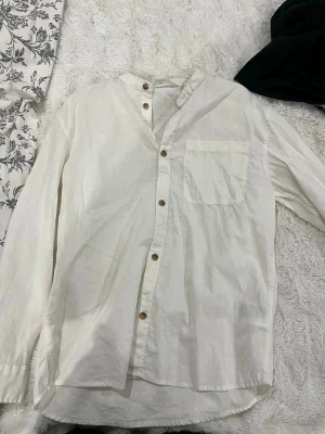 Vit henley-skjorta från H&M - Säljer en stilren vit henley-skjorta från H&M i storlek 152. Skjortan har lång ärm, bröstficka och träknappar framtill. Tillverkad i ett lätt och luftigt linnematerial, perfekt för en clean och avslappnad look.