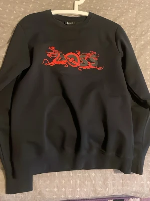 Svart sweatshirt från BOSS med drakmotiv - Svart sweatshirt från BOSS i storlek S med ett fett rött drakmotiv och BOSS-logga på bröstet. Tröjan har rund halsringning och långa ärmar. Perfekt för dig som vill sticka ut med en unik detalj på din outfit.