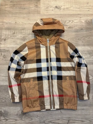 Burberry - Säljer en klassisk rutig vindjacka från Burberry i beige, svart, vit och röd. Jackan har huva med snörning, dragkedja framtill och två fickor. Tillverkad i polyester och har den ikoniska Burberry-rutan. Perfekt för dig som vill sticka ut med stil. Storlek: XL junior så passar M män