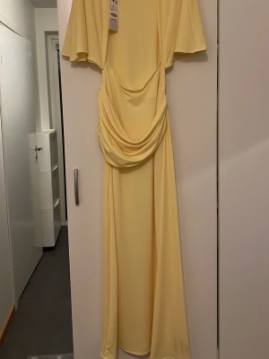 Ljusgul långklänning med drapering - Säljer en elegant ljusgul långklänning med vackra draperingar framtill och vida korta ärmar. Klänningen har en mjuk, flytande form och är perfekt för sommarens speciella tillfällen. Tillverkad i ett lätt och följsamt material som ger en lyxig känsla.