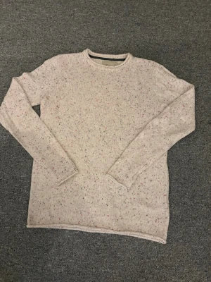 Beige stickad tröja från Revolution - Säljer en beige stickad tröja från Revolution i storlek M. Tröjan har ett diskret prickigt mönster i rött och svart, rund halsringning och långa ärmar. Perfekt för dig som gillar enkel men snygg stil.