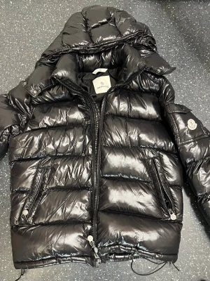 Svart Moncler pufferjacka med luva - Säljer en svart pufferjacka från moncler den är glansig utsidan så finns de två fickor med drag kedja sen i armen finns de moncler loggan där man kan skanna och en knapp som man kan lägga saker i insidan så finns de också en till ficka prisen kan diskuteras 