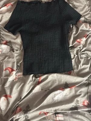 Svart ribbad topp från H&M - Säljer en svart ribbad topp från H&M med offshoulder-design och korta ärmar. Tyget är stretchigt och har en smockad struktur som ger en snygg passform. Perfekt till jeans eller kjol för en trendig look.