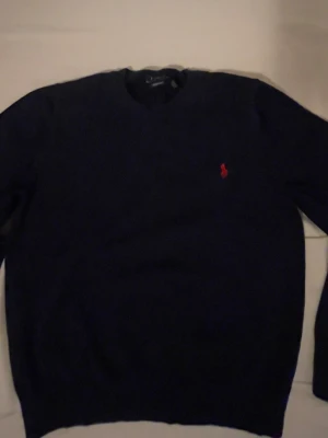 Marinblå stickad tröja från Ralph Lauren - Klassisk marinblå stickad tröja från Ralph Lauren med rund halsringning och den ikoniska röda loggan broderad på bröstet. Tröjan har långa ärmar och ribbade muddar vid hals, ärmslut och nederkant. Perfekt till lager på lager under höst och vinter.PRIS GÅR ATT DISKUTERA
