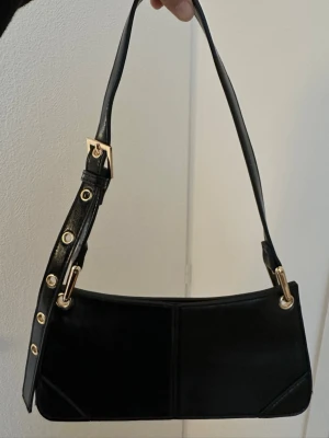 Svart axelväska i skinn med gulddetaljer - Säljer en stilren svart axelväska i skinn med justerbart axelband och snygga guldfärgade metalldetaljer. Väskan har en avlång, rektangulär form och minimalistisk design som passar till många olika looks.