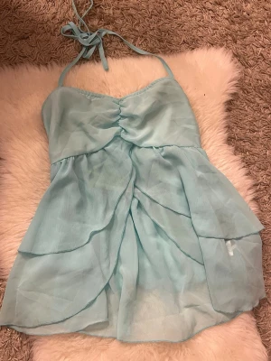 Ljusblå omlott topp med volanger - Supersöt ljusblå topp med tunna axelband och omlottdetaljer framtill. Toppen har flera lager av volanger och ett lätt, luftigt tyg som ger en romantisk känsla. Perfekt för sommarens alla tillfällen!