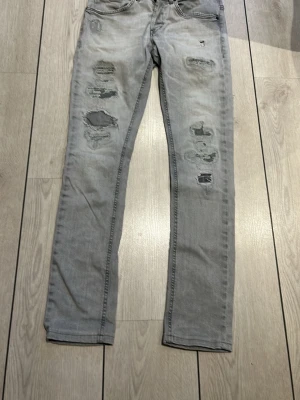 Grå dondup - Säljer ett par grå skinny jeans med slitningar och hål på framsidan. Jeansen har klassisk femficksdesign och är i stretchigt denimtyg för skön passform. Perfekta för dig som gillar en edgy och avslappnad stil.