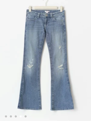 Blå low waist flare jeans med paljettdetalj - Säljer ett par ljusblå jeans med låg midja och utsvängda ben. Jeansen har slitna detaljer framtill och bakfickor med broderi och silvriga paljetter. Perfekta för dig som gillar lite extra bling på dina jeans! Helt oanvända prislappar sitter kvar.