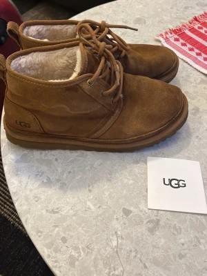  UGG boots i mocka med snörning - Säljer ett par klassiska bruna UGG boots i mjuk mocka med snörning och fluffigt vitt foder. Skorna har rund tå, platt sula och är perfekta för kalla dagar. UGG-logga på sidan och slitstark yttersula. Kommer med originalkartong.