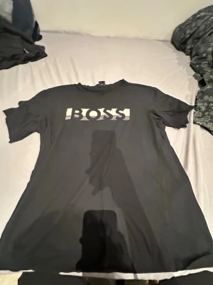 Svart BOSS t-shirt med tryck XL - Svart t-shirt från BOSS i storlek XL med stort vitt BOSS-tryck på bröstet. Klassisk rund hals och korta ärmar. Materialet är mjuk bomull som känns skönt mot huden. Perfekt för dig som gillar stilrena och enkla plagg med tydlig logga.