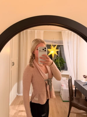 Rosa kofta från Odd Molly med grå knytrosett - Supersöt ljusrosa kofta från Odd Molly med diskret mönster och grå knytrosett framtill. Koftan har lång ärm, peplumkant och är stickad i mjukt material. Koftan är i väldigt bra skick. Ha i åtanke att den är för liten för mig på bilden 