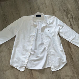 Vit skjorta från Ralph Lauren - Klassisk vit skjorta från Ralph Lauren med broderad logga på bröstet. Skjortan har lång ärm, knappar framtill och en snygg krage. Tillverkad i mjuk bomull som känns skön mot huden. Perfekt för dig som gillar stilrena och tidlösa plagg. Pris går att diskuteras. 