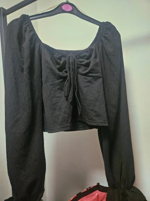 Svart blus med snörning - Trendig svart blus med långa puffärmar och snygg snörning framtill. Toppen har en fyrkantig ringning och är gjord i ett lätt, krinklat tyg som ger en cool struktur. Bra stretch pga smycken och använd en eller två ggr.