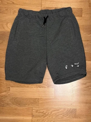 Mörkgrå shorts XXL - Säljer ett par mörkgrå shorts  i storlek XXL. De har elastisk midja med svart snörning, två sidofickor och vit broderad Off-White-logga framtill. Tillverkade i mjuk bomull, perfekta för chill dagar eller träning.