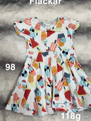 Färgglad klänning med glassmönster - Supersöt klänning från Polarn o pyret med färgglatt glass- och fruktmönster på vit botten. Klänningen har korta ärmar och en snurrig kjol, perfekt för sommarens alla äventyr. 🌸 Fläckar. Storlek 98