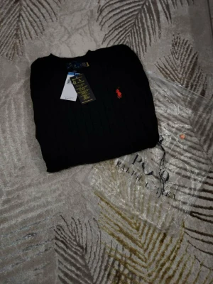 Svart stickad tröja Polo Ralph Lauren - Säljer en svart stickad tröja från Polo Ralph Lauren i storlek M. Tröjan har ett klassiskt kabelstickat mönster och den ikoniska röda loggan broderad på bröstet. Rund halsringning och långa ärmar. Perfekt för dig som vill ha en stilren och tidlös look.
