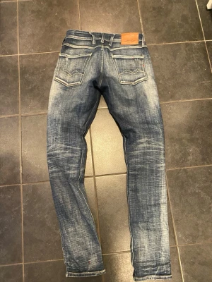 Replay Anbass blå slitna jeans - Säljer ett par Replay Anbass jeans i blå tvätt med slitningar och distressed detaljer. Jeansen har smal passform, klassiska fem fickor och snygga kontrastsömmar. Märkeslapp bak i midjan och Replay-logga vid fickan. Perfekta för en avslappnad streetstil.