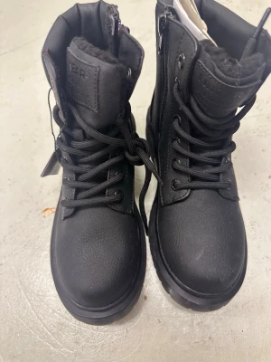 Svarta boots från Linear of Sweden - Tuffa svarta boots från Linear of Sweden med grov sula och snörning. Skorna har dragkedja på sidan för enkel påtagning och är fodrade med mjukt, varmt foder. Tillverkade i syntetmaterial med rund tå och robust design, perfekta för kalla dagar.