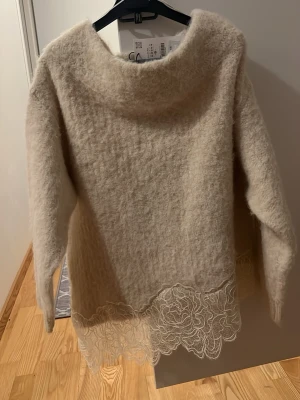 Beige fluffig stickad tröja med spets - Superfin beige stickad tröja med fluffig känsla och bred halsringning. Tröjan har långa ärmar och en unik detalj med spets längst ner som ger en romantisk touch. Perfekt för dig som vill ha något mysigt men ändå extra.