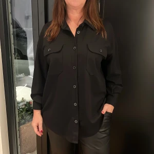 Svart oversized skjorta med fickor - Säljer en svart oversized skjorta med stora bröstfickor och knappar framtill. Skjortan har lång ärm och en klassisk krage, perfekt för en clean och stilren look. Materialet känns mjukt och följsamt. Storlek L, stor i storleken. 