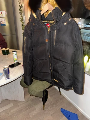 Svart Moose Knuckles pufferjacka XXL - Säljer en svart pufferjacka från Moose Knuckles i storlek XXL. Jackan har guldfärgade detaljer, dragkedjor och knappar, samt en stor svart pälskantad huva. Fodret är guldigt med Moose Knuckles-logga. Perfekt för kalla dagar. Saknar kvitto därav priset 
