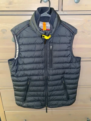 Svart dunväst från Parajumpers XS - Snygg svart dunväst från Parajumpers i superlätt material. Västen har hög krage, dragkedja framtill och tre fickor med dragkedja. Detaljer i gult och orange vid halsen ger en sportig touch. Perfekt att slänga över en hoodie eller tröja.