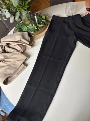 Svarta kostymbyxor från H&M, strl 38 - Säljer ett par klassiska svarta kostymbyxor från H&M i storlek 38. Byxorna har rak passform, hög midja och är tillverkade i ett mjukt, vävt material. Perfekta för en clean och stilren look. Diskreta fickor i sidorna och hällor för bälte. Short modell 