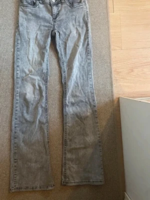 Grå bootcut jeans från Nelly - Säljer ett par grå bootcut jeans från Nelly med snygg tvättad look och svarta broderade detaljer på bakfickorna. Jeansen har hög midja, dubbla knappar och klassisk femficksdesign. Materialet är jeans med en mjuk känsla och passformen är loose