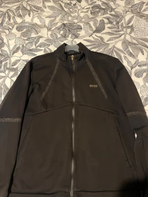 Svart zip hoodie från BOSS - Säljer en svart hoodie från BOSS med hel dragkedja och guldfärgade detaljer. Tröjan har diskreta mönstrade band längs axlar och ärmar samt BOSS-logga på bröstet. Tillverkad i mjuk bomullsmix och har två fickor framtill. Perfekt för en clean och stilren look.