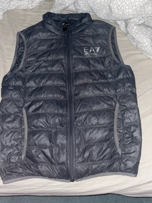 Mörkgrå dunväst från Emporio Armani - Säljer en stilren mörkgrå dunväst från Emporio Armani EA7 pga att den är för liten . Västen har quiltad design, hög krage och dragkedja framtill. Två fickor på sidorna och diskret logga på bröstet. Perfekt för lager-på-lager och snygg till både hoodie och t-shirt. Använd 1 sommar sen har den bara hängt.
