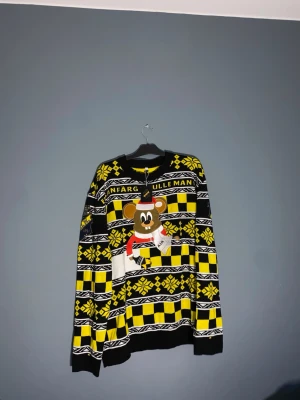 AIK jultröja XXL med musmotiv - Säljer en stickad AIK jultröja i svart, gul och vit med stort musmotiv på bröstet och texten 'FÄRGMAN'. Tröjan har klassiskt vintermönster, lång ärm och ribbade muddar. Perfekt för dig som vill sticka ut på julmyset!