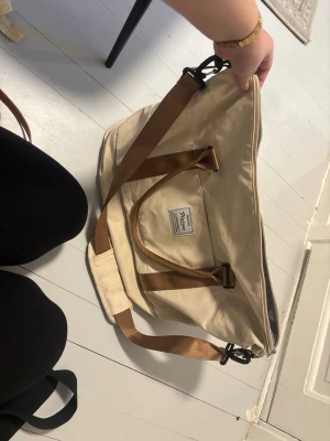 Beige weekendbag från Nilson - Snygg beige weekendbag från Nilson med bruna axelband och detaljer. Väskan har ett stort huvudfack med ljusgrått innerfoder och dragkedja. Perfekt storlek för övernattning eller gym och enkel att bära med både handtag och axelrem.