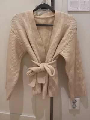 Beige ribbad omlottkofta med knytband - Superfin beige ribbad omlottkofta med knytband i midjan. Koftan har långa ärmar och är gjord i ett mjukt stickat material som känns både cozy och trendigt. Perfekt att slänga på sig över ett linne eller t-shirt för en chill look.