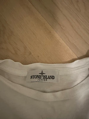 Stine Island T-Shirt - Vit t-shirt från Stone Island Junior med tryck på framsidan. Klassisk rund hals och korta ärmar. Materialet är mjuk bomull och loggan syns både på ryggen och i trycket framtill. Perfekt för sommarn.