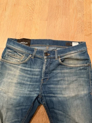 Blå jeans från Dondup, storlek 31 - Säljer ett par klassiska blå jeans från Dondup i storlek 31. Jeansen har en snygg tvätt med slitningar, fem fickor och raka ben. Tillverkade i mjukt jeanstyg med kontrastsömmar och detaljerade bakfickor. Perfekta för dig som gillar en tidlös och avslappnad look.