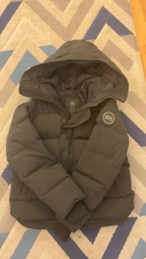  Canada Goose jacka - Svart pufferjacka från Canada Goose med stor huva och ikonisk patch på ärmen. Jackan har dragkedja och knappar framtill, samt quiltad design som ger extra värme. Perfekt för kalla dagar och har en clean, stilren look.