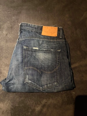 Jack & Jones jeans  - Säljer ett par blå jeans från Jack & Jones med slitningar och distressed detaljer. Klassisk femficksmodell med raka ben och normal passform. Jeansen har en snygg tvättad look och patch i läderimitation bak i midjan. Perfekta för en avslappnad stil.