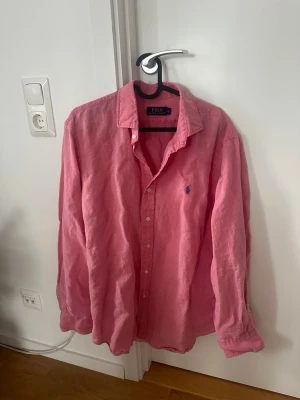 Rosa linneskjorta från Polo Ralph Lauren - Säljer en snygg rosa skjorta från Polo Ralph Lauren i storlek L. Skjortan är tillverkad i linne, har långa ärmar, klassisk krage och knäppning framtill. Den ikoniska loggan är broderad på bröstet. Perfekt för dig som gillar stilrena och bekväma plagg.