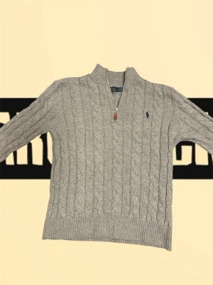 Grå quarter zip från Polo Ralph Lauren - Säljer en grå kabelstickad tröja från Polo Ralph Lauren med half zip och hög krage. Tröjan har det klassiska mörkblå logobroderiet på bröstet och ribbade muddar. Perfekt för dig som vill ha en stilren och bekväm look.