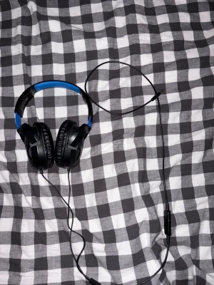 Kabelanslutna over-ear hörlurar - Bekväma over-ear hörlurar med kabelanslutning och vadderad bygel. Svart och blå design, mjuka öronkuddar och inbyggd mikrofon på sladden. Passar bra för gaming, musik och samtal. Ser ut att vara i gott skick utan synligt slitage.