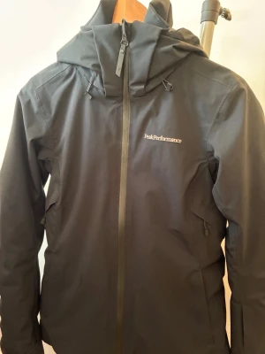 Svart skidjacka från Peak Performance XS - Säljer en svart skidjacka från Peak Performance med huva och hel dragkedja framtill. Jackan har diskret logga på bröstet och är tillverkad i slitstarkt syntetmaterial. Två sidofickor med dragkedja och justerbar huva för extra funktionalitet.