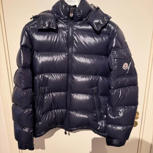 Moncler maya size 3 - Säljer en mörkblå dunjacka från Moncler med glansig finish och avtagbar huva. Jackan har två sidofickor med dragkedja, ikonisk Moncler-logga på ärmen och coola detaljer på dragkedjan. Perfekt för kalla dagar och har en schysst, puffad look.