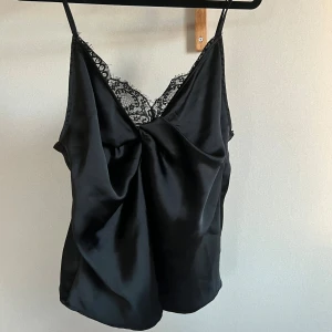 Svart satin linnetopp med spets - Snygg svart linnetopp i satin med tunna axelband och v-ringad hals med dekorativ svart spets. 
