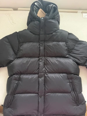 Svart pufferjacka Burberry XXL - Svart pufferjacka från Burberry i storlek XXL med avtagbara ärmar så den kan användas som väst. Jackan har en stor huva fodrad med grått och vitt rutigt tyg, dragkedja och tryckknappar framtill samt två sidofickor med dragkedja. Perfekt för kalla dagar.
