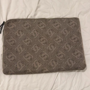 Beige datorväska med mönster - 14 tum. Stilren beige datorväska med diskret mönster av svarta loggor i rutigt utförande. Väskan har dragkedja upptill och är perfekt för att bära din laptop eller viktiga papper. Materialet känns tåligt och har en snygg struktur. Har någon fläck därav priset.